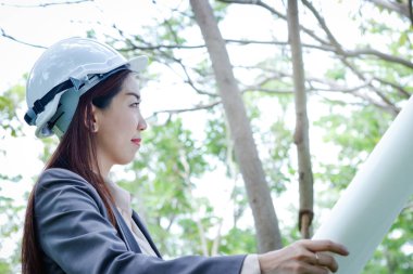 Kadın çevre mühendisi, takım elbise giyiyor, beyaz şapka takıyor, elinde kağıt bir plan tutuyor.