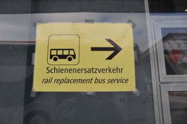Sarı demiryolu servis tabelası, siyah harfler ve piktogram, İngilizce ve Almanca (Schienenersatzverkehr)