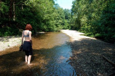 Yazın nehirde çıplak ayakla yürüyen bir kadın, ağaçlarla çevrili bir nehir.