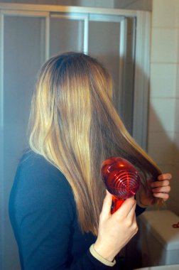 Genç bir kadın uzun sarı saçlarını kızıl saç kurutma makinesiyle kurutuyor.
