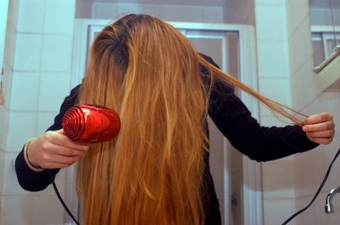 Genç bir kadın uzun sarı saçlarını kızıl saç kurutma makinesiyle kurutuyor.