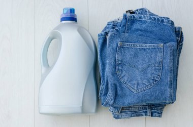 Masanın üstünde büyük plastik bir şişe sıvı toz ve bir yığın kot eşya var. Deterjanlar ve temizlik ürünleri.