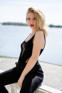 Güzel, genç bir model. Sarışın doğada poz veriyor. Dudaklarında kırmızı ruj var. Siyah cisim içinde model