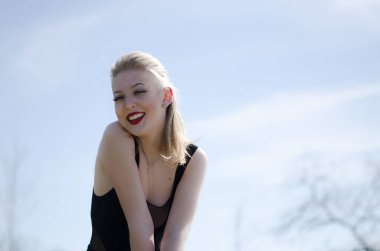 Güzel, genç bir model. Sarışın doğada poz veriyor. Dudaklarında kırmızı ruj var. Siyah cisim içinde model