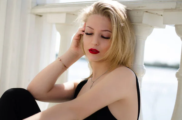 Young beautiful girl. Blonde posing while sitting by the column. Red lipstick on the lips. Photo of a model in nature