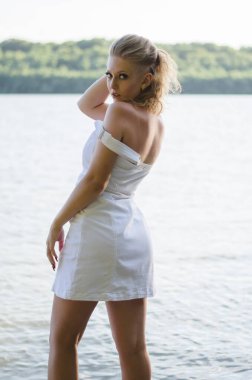 Güzel sarışın bir kız nehir kıyısında duruyor. Doğada poz veren beyaz elbiseli bir model.