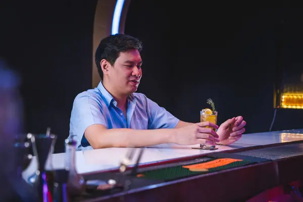 Asyalı erkek, gece kulübü gişesinde yalnız başına arkadaşlarını bekler. Mavi gömlekli, viski içen, yalnız başına içki içen, bar restoranı, mankenler çerçevenin ortasına oturur.