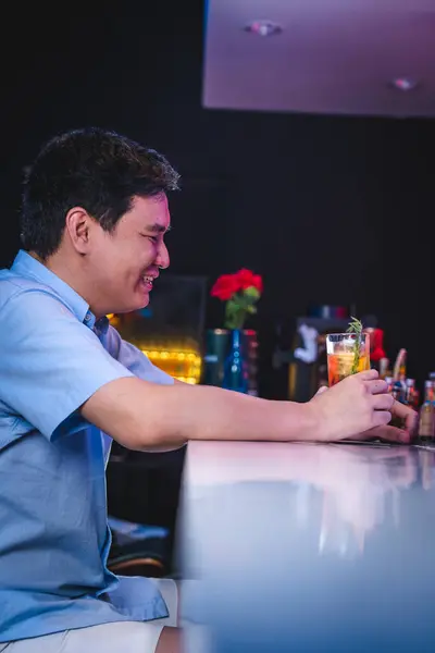 Asyalı bir adam içki barında tek başına otururken gülümsüyor, mavi tişörtlü bir adam oturuyor, içiyor, içki içiyor, bir bardak alkolle eğleniyor, erkek yalnız içiyor, bar restoranında.