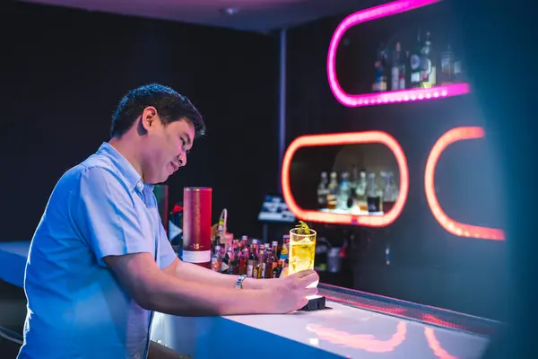 Asian man at whiskey beverage bar in colorful nightclub, male wear blue shirt night out, purple orange decoration light on background, male drinking alone, whiskey vodka bottles collection, classy man