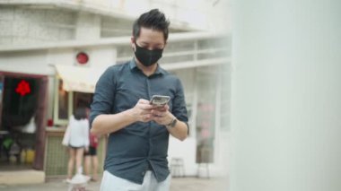 Asyalı adam şehirde akıllı telefon kullanarak maske takıyor.