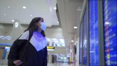 Asyalı kadın transfer yolcusu koruyucu maske takıyor kalkış zamanı masa ekranına bakıyor, havaalanı terminalinde, yurtiçi covid-19 salgınından ayrılıyor, yeni normal uçuş zamanı