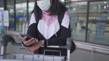 Korumalı maskeli bir kadın havaalanında tramvay bagajlı akıllı telefon kullanıyor. Corona virüsü salgını, yeni normal sosyal mesafe, havaalanı terminalinin dışında bekleyen kadın.