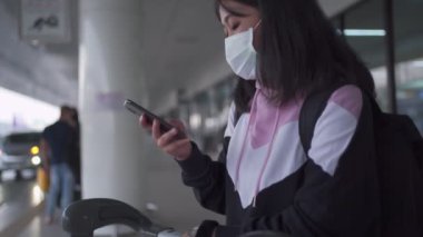 Koruyucu maskeli Asyalı bir kadın havaalanında arka planında bagaj arabasıyla akıllı telefon kullanıyor. Salgın hastalık salgını var. Yeni normal sosyal mesafe.