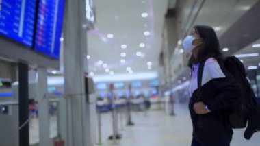 Asyalı kadın transfer yolcusu koruyucu maske takıyor kalkış zamanı masa ekranına bakıyor, havaalanı terminalinde, yurtiçi covid-19 salgınından ayrılıyor, yeni normal uçuş zamanı