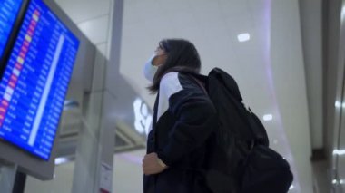 Asyalı kadın transfer yolcusu koruyucu maske takıyor kalkış zamanı masa ekranına bakıyor, havaalanı terminalinde, yurtiçi covid-19 salgınından ayrılıyor, yeni normal uçuş zamanı  