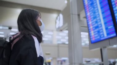 Beyaz maskeli Asyalı kadın havaalanı uçuş saatlerine bakıyor, havaalanı terminalinde, Corona virüsü covid-19 salgını, yeni normal yaşam tarzı sosyal mesafe, mavi ekran şehri terk ediyor, 4K