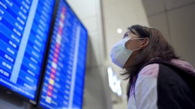 Asyalı kadın transfer yolcusu koruyucu maske takıyor kalkış zamanı masa ekranına bakıyor, havaalanı terminalinde, yurtiçi covid-19 salgınından ayrılıyor, yeni normal uçuş zamanı   