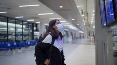 Asyalı kadın transfer yolcusu koruyucu maske takıyor kalkış zamanı masa ekranına bakıyor, havaalanı terminalinde, yurtiçi covid-19 salgınından ayrılıyor, yeni normal uçuş zamanı   