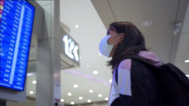 Asyalı kadın transfer yolcusu koruyucu maske takıyor kalkış zamanı masa ekranına bakıyor, havaalanı terminalinde, yurtiçi covid-19 salgınından ayrılıyor, yeni normal uçuş zamanı   