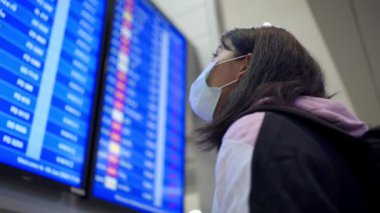Asyalı kadın transfer yolcusu koruyucu maske takıyor kalkış zamanı masa ekranına bakıyor, havaalanı terminalinde, yurtiçi covid-19 salgınından ayrılıyor, yeni normal uçuş zamanı   