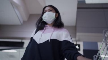 Beyaz maskeli Asyalı bir kadın alışveriş merkezinde yürüyen merdivenden iniyor, salgın hastalık salgını, yeni normal sosyal mesafe, maskeli bir kadın yürüyen merdivenden iniyor, bina içinde, halka açık bir yerde.