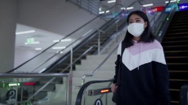 Asyalı kadın koruyucu maske takıyor havaalanı seyahatinde yürüyen merdivenden iniyor, covid-19 salgını, yeni normal sosyal mesafe, yürüyen merdivenden iniyor, kapalı bina, sosyal güvenlik merdivenleri.