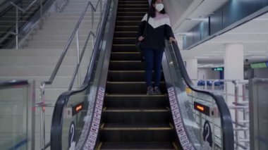 Beyaz maskeli Asyalı bir kadın alışveriş merkezinde yürüyen merdivenden iniyor, salgın hastalık salgını, yeni normal sosyal mesafe, maskeli bir kadın yürüyen merdivenden iniyor, bina içinde, halka açık bir yerde.