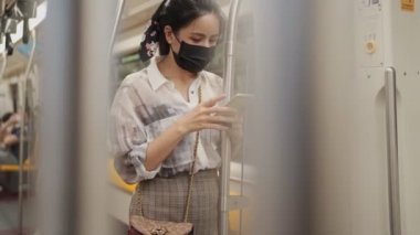 Güzel bir Asyalı kadın siyah maske takıyor metro vagonunda direğe tutunuyor cep telefonu kullanıyor, banliyö yolunda, yeni normal, toplu taşıma riski, sosyal mesafe, akıllı telefon kullanıcısı...
