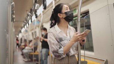 Siyah koruyucu maskeli, akıllı telefon kullanan Asyalı bir kadın, direğe yaslanmış, yeni normal, metroda kendini koruyan bir kadın, toplu taşıma araçları, sosyal mesafeler, merak içinde kaybolmuş. 