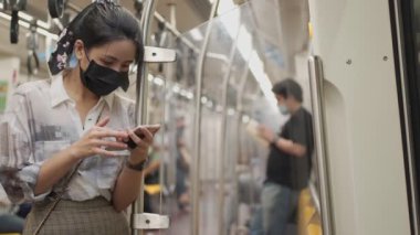 Siyah koruyucu maskeli bir Asyalı kadın akıllı telefon kullanıyor, direğe yaslanıyor, yeni normal, metro metrosunda kadın, toplu taşımada kendini koruma, sosyal mesafeler, ağ bağlantısı.