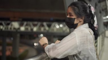 Güzel Asyalı kız köprüden karşıya geçerken siyah koruyucu maske takıyor. Rahatlama, dikkat çekme, arka planda yürüme, sosyal mesafeler, gece vakti.