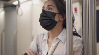 Asyalı genç bir kadın siyah koruyucu maske takıyor, metro istasyonundaki zaman çizelgesine bakıyor, yeni normal sosyal uzaklık yaşam tarzı, kişisel koruma, boş metro vagonu, bulaşıcı risk uyarısı yavaş hareket