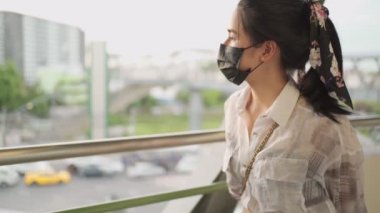 Genç Asyalı bir kadın siyah koruyucu bir maske takıyor ve şehir trafiğine bakıyor, gökyüzü köprüsü yolu boyunca, genç bir kadın yürümeye devam ediyor sokak arkası, altın gün batımı trafiği
