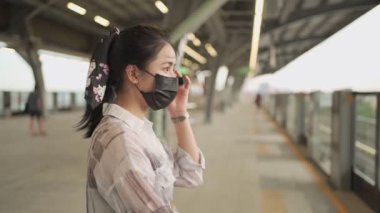 Asyalı kadın banliyö görevlisi siyah maske takıyor gökyüzü tren platformunda duruyor, covid-19 salgını, metro istasyonunda yalnız umutsuz bir kadın. Yeni normal, ayar maskesi, toplu taşıma, gün ışığı, yavaş çekim.