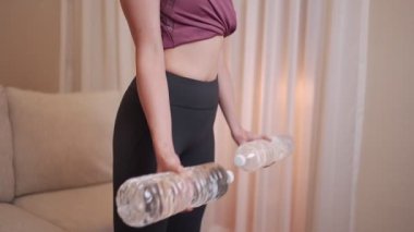 Genç, formda bir kadın oturma odasında egzersiz yapıyor. Plastik su şişesi silah egzersizi, biseps kıvrım dayanıklılık tekrarlama eğitimi, covid-19 karantinası sırasında sağlıklı yaşam tarzı.