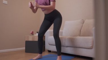 Evde yoga minderine çömelmiş bir kadın, oturma odasında ayakta egzersiz yapıyor, ayak egzersizi yapıyor, ev bacakları egzersizi yapıyor, kovid-19 karantinası sırasında sağlıklı yaşam tarzı, kilo kaybı, vücut ağırlığı egzersizi, arka planda kanepe.