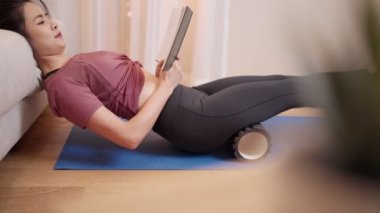 Genç Asyalı fit ve sıkı kadın kitap okuyor ve oturma odasında yoga matt üzerinde köpük kullanıyor. Boş zaman, sağlık hizmetleri kişisel gelişim, vücut geliştirme, modern kadın sağlıklı yaşam tarzı.