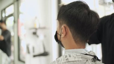 Berber dükkanının arkasından manzara, siyah maskeli Asyalı adam, ayna yansımalı saç spreyi, keskin kenarlı favoriler, sabah tımarı, su spreyi, salgından sonra Covid-19 yeniden açılan iş yeri.