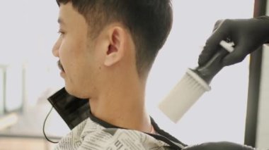 Berber, saç kesiminden sonra müşteriyi temizlemek için fırça kullanır, genç adam saçını kestirir, erkek berber dükkanı, kuaför dükkanı, kuaför mesleği fırsatı, yeniden iş imkanı, yan görüş, doğal ışık.