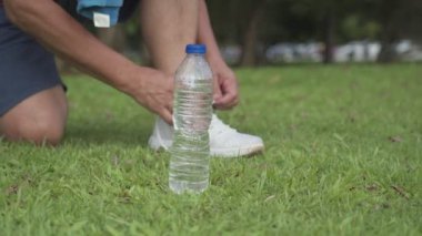Asyalı bir adam ayakkabılarını bağlıyor yeşil çimlerde koşu yaparken spor yaparken, açık havada egzersiz yaparken, aktif yaşam tarzında, susuz kalmadan, su şişesini yan tarafa yerleştirirken, koşucu su şişesini kapıp