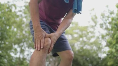 Asyalı tenli erkek, spor yaparken dizinden yaralı. Arka planda ağaçlarla birlikte parkta koşuyor. Vücut durumu, diz ağrısı, eklem bağı sorunu, dışarıda diz ağrısı. 