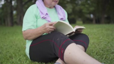 Asyalı son sınıf öğrencisi kadın spor sonrası parkta oturup kitap okuyor. Yaşlılık aktivitesi, emeklilik hayatı, yaşam enerjisi rahatlama, rahat çimenlerin üzerinde obez kadın okuyucu. 