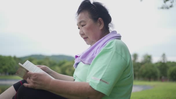 アジアのシニア女性は 運動後 屋外公園で休憩と読書本を座って 古い年齢レジャー活動 退職生活 ウェルネス活力リラクゼーション 快適な草の上の肥満の女性リーダー — ストック動画
