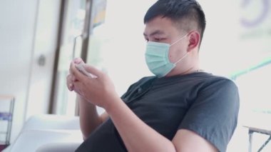 Asyalı adam, Coronavirus salgını sırasında doktorla görüşmek için beklerken cerrahi maske takıyor. Telefon rezervasyonu yapan, kablosuz kolaylık teknolojisi kullanan hasta.   