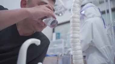 Asyalı erkek hasta diş sağlığı kliniğinde ağzını çalkalayıp duruyor, tıbbi ağız kontrolü yapıyor, sıhhiye kıyafeti giyiyor, covid-19 Corona virüsü sırasında, konsültasyon kontrolü yapıyor, bakterileri öldürüyor.