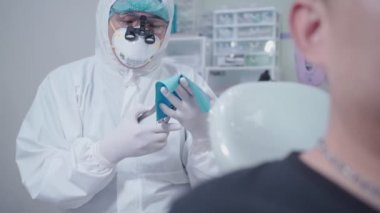 Ameliyattan önce kauçuk baraj hazırlayan tam donanımlı bir erkek dişçi, diş prosedürü odası, beyaz PPE giysisi, tıbbi cerrahi ekipman, dezenfekte edilmiş tesis, koronavirüs yayılması önleme, kişisel ağız temizliği