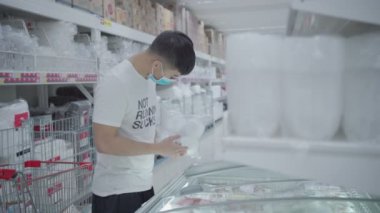 Genç Asyalı bir adam cam sürgülü kapıyı açar, süpermarketin dondurucusundan dondurulmuş yiyecek paketleri alır, bir tane alır, tasarruf fiyatı, gıda endüstrisinde teknoloji, salgın sırasında stok yapar.  