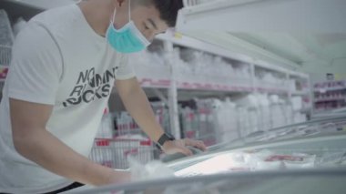 Doğu Asyalı bir adamın süpermarketin dondurucusu için dondurulmuş yiyecek almaya karar verirken, el aynasından buzdolabının kapağını yavaşça açarken, covid-19 salgını sırasında tıbbi maske takarken, yiyecek seçerken,