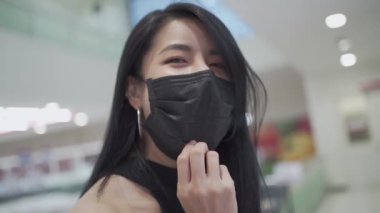 Kovid-19 koronavirüs salgını sırasında marketin içinde yürürken genç çekici Asyalı kadının maskesinin altında yavaşça gülümsemek, karantina mesafesini korumak, kameraya bakan model