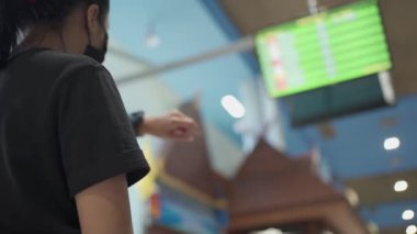 Arkadaki genç kadın maske takıyor. Havaalanı zaman tablosu, terminal kalkış platformu, toplu taşıma, yatış saati, covid-19 salgını, yeni normal sosyal mesafe, son çağrı. 